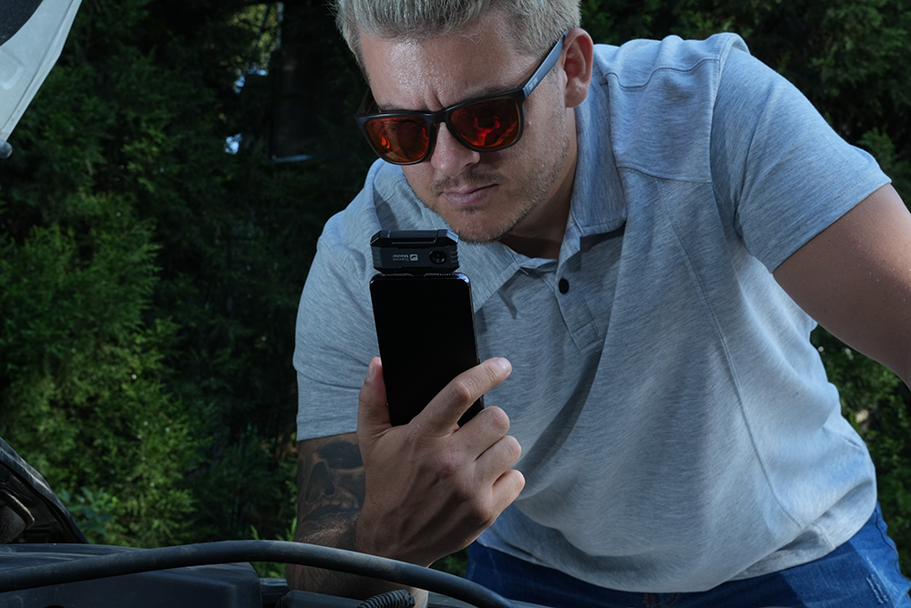 Man using a Thermal Master P1 smartphone thermal camera to inspect a car engine for heat-related issues.