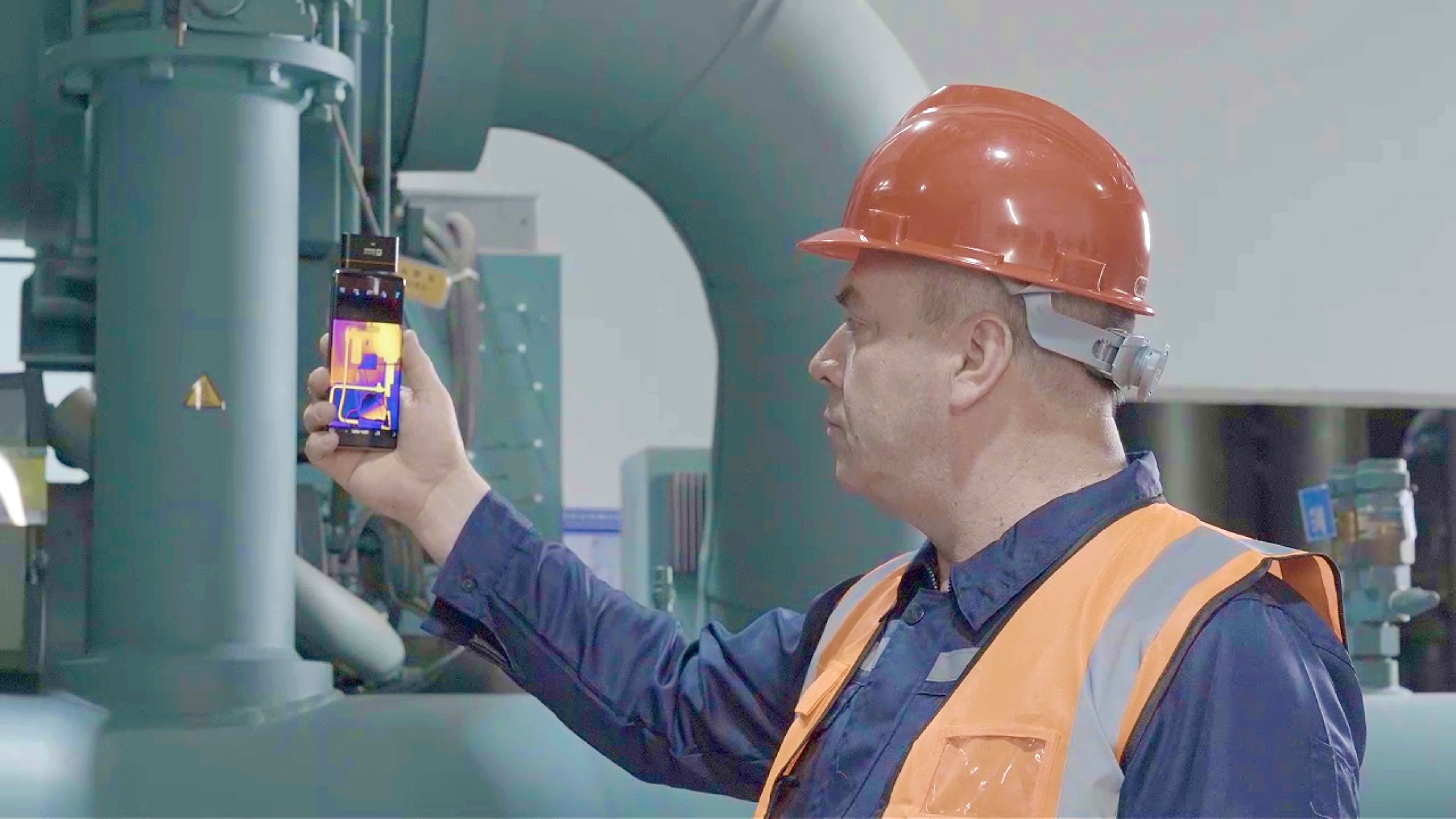 Industrial technician using a smartphone thermal camera to inspect machinery and detect heat anomalies.